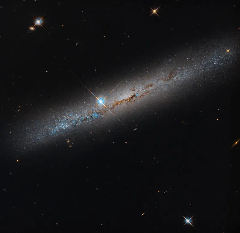 A long, pale streak of a galaxy slanting across a black field from lower left to upper right, threaded with reddish-brown dust and blue knots; left of the galaxy's centre, a bright star shows four spikes.