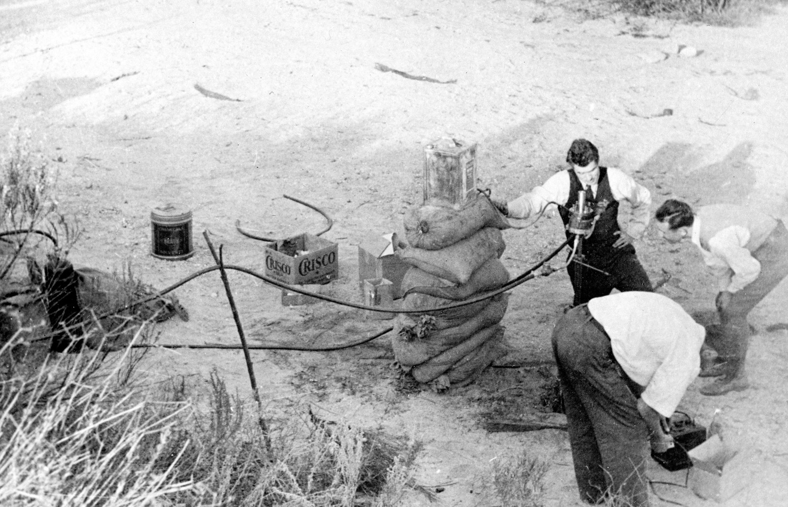 Photo en noir et blanc : trois hommes s’affairent autour d’un dispositif d’essai, sur un sol sablonneux, à côté d’une haute pile de sacs de sable. L’un, en gilet sombre, se tient près du moteur ; les deux autres, en chemise blanche, sont penchés sur l’appareillage. Des tuyaux, des bidons métalliques et un carton marqué « Crisco » traînent autour d’eux.
