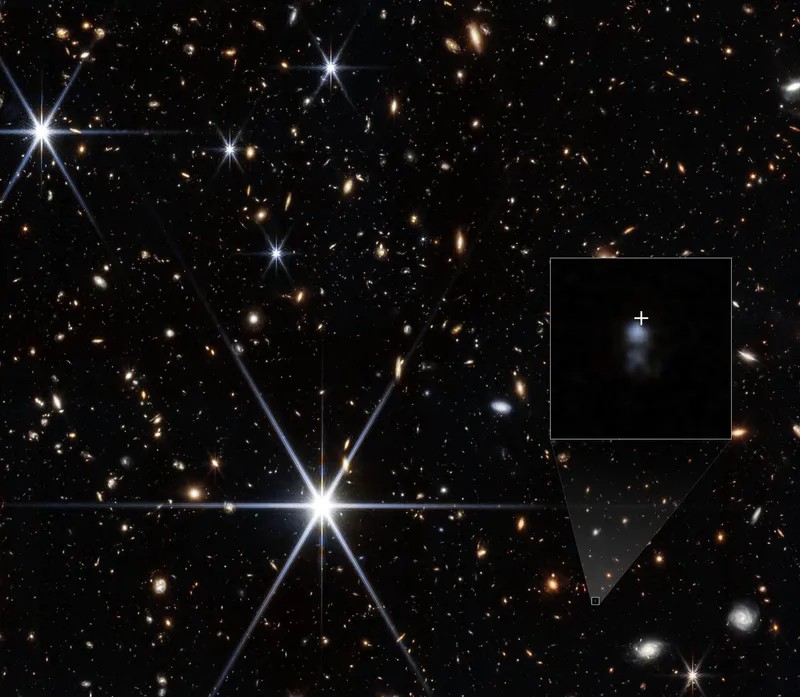 A dark field crowded with small, mostly orange galaxies and a few bright stars with long spikes. A box at lower right is enlarged in an inset showing two faint blue smudges, with a white cross just above the upper one.