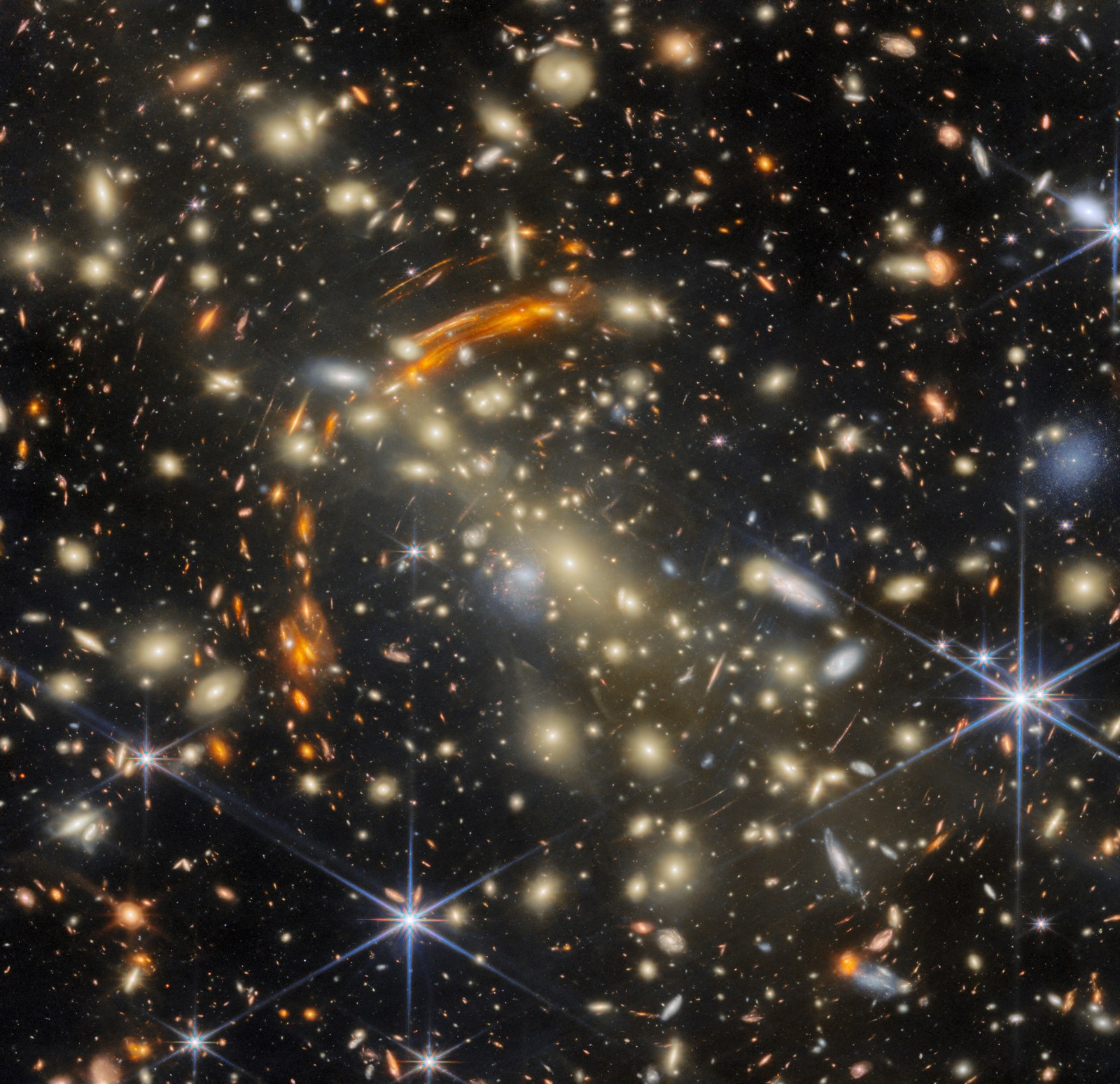 Ett tätt fält av gyllene elliptiska galaxer med en ljus galax nära mitten. Böjda orange bågar löper längs hopens övre vänstra del och vänstra sida, och till höger och nedtill syns ljusa förgrundsstjärnor med långa blå strålar.